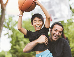A father holds a boy with a basketball on an outdoor basketball court.