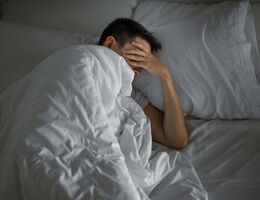 A man huddles under his covers in bed with a hand over his eyes.