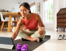 A woman sits on an exercise mat and reaches for her tablet.