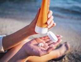 Hands squeeze sunscreen from a tube at a beach.